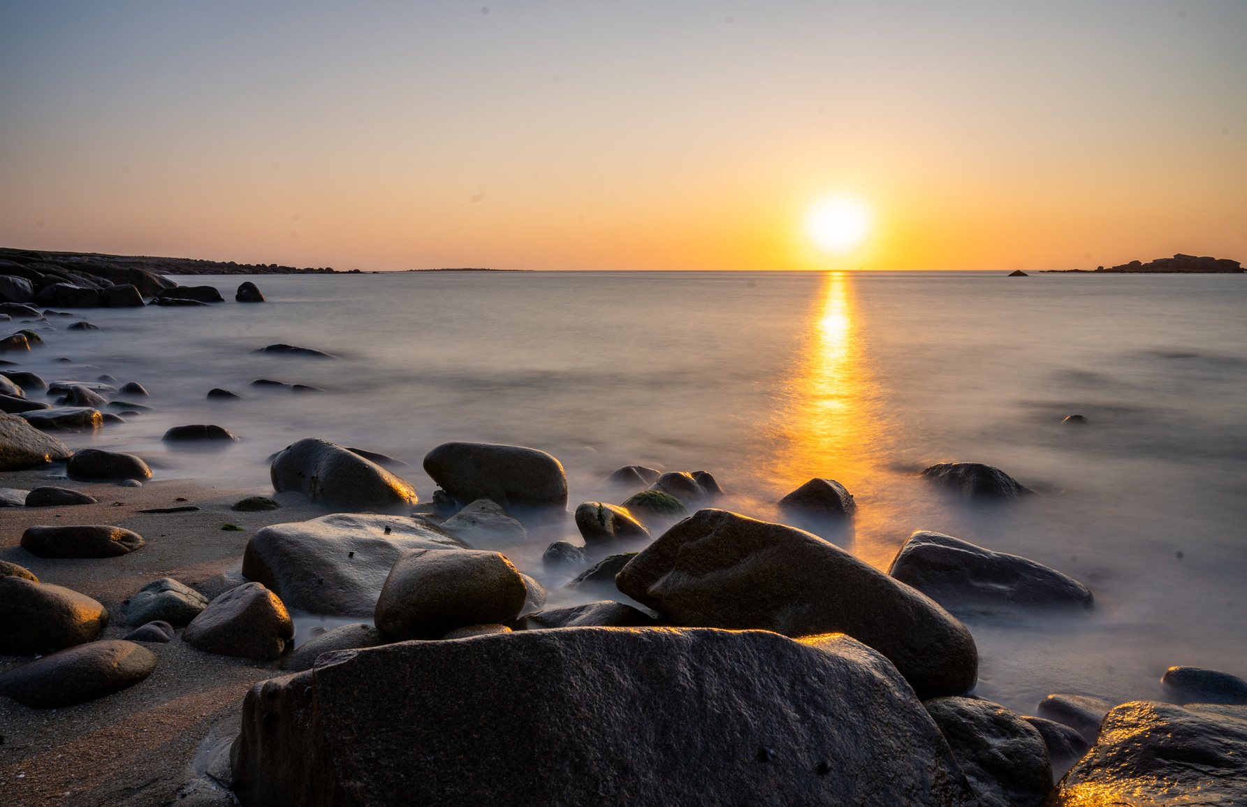 Photographie d’un coucher de soleil sur une plage rocheuse. La lumière dorée du soleil se reflète sur l’eau calme, contrastant avec les rochers sombres en premier plan. Une longue exposition donne un effet soyeux à la mer.