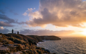 Vue du Cap Fréhel en Bretagne au coucher du soleil, avec le phare illuminé par une lumière dorée, les falaises plongeant dans l’océan et de grandes formations nuageuses dans le ciel.
