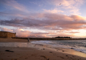 Deux personnes pêchent sur une plage au crépuscule, sous un ciel pastel rempli de nuages dorés et rosés, avec les remparts d’une ville en arrière-plan.