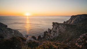Coucher de soleil sur la mer vu depuis les falaises rocheuses d’Erquy, en Bretagne, avec une lumière dorée se reflétant sur l’eau calme.