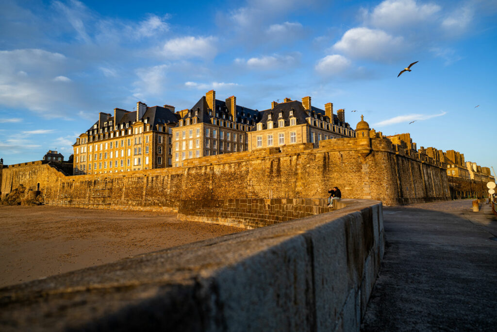 Vue des remparts de Saint-Malo au coucher du soleil, avec des façades de granit éclairées par une lumière dorée. Un couple est assis sur le bord du chemin, et des mouettes volent dans le ciel bleu.